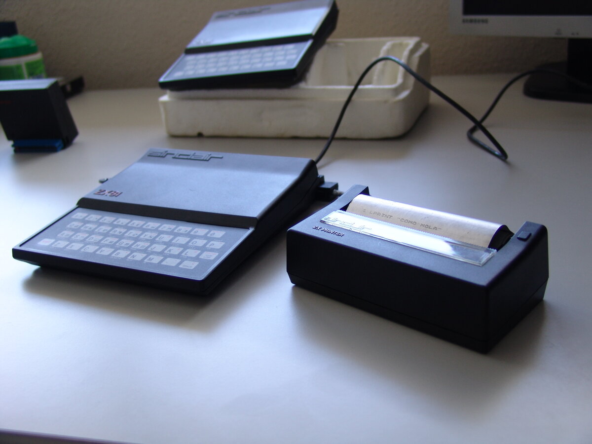 A ZX81 connected via cable to a Sinclair ZX Printer on a desk, mid-print, with a curl of shiny metallic paper emerging showing the printed line 1 LPRINT COMO HOLA